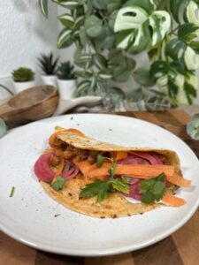 Veganer Taco mit Jackfruit und Kichererbsen, grüner Salsa und frischen Zutaten angerichtet auf einem Teller auf einem Holzbrett